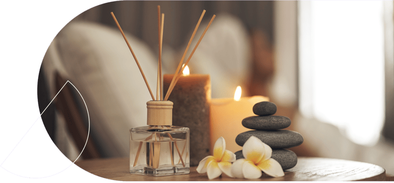 perfume diffuser, 2 candles, 4 grey pebbles, and two flowers on a table with a sofa in the background